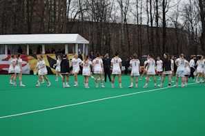 A group of female athletes in white sports uniforms are positioned on a green sports field. The players are wearing white shorts and jerseys with numbers on their backs, accompanied by sports shoes. They appear to be gathering in a somewhat circular formation, possibly preparing for a game or practice. Behind them, there is a white tent with several people inside, and a forested area with leafless trees, suggesting it is a cool season.
