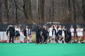 A group of athletes in white sports uniforms are gathered on a green field, holding lacrosse sticks. They appear to be part of a team, engaging in conversation and organizing equipment. The background shows a forested area with bare trees and a dark fence.