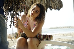 A woman in a pink and white striped bikini is seated on a beach chair under a thatched umbrella on a sandy beach. She is examining a collection of various colorful beaded necklaces and bracelets displayed by a vendor. The background shows the sea and a rocky breakwater.