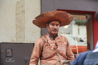 A person dressed as a cowboy statue, fully painted in a copper or bronze color, wearing a large brim hat, a bow tie, and traditional cowboy attire. The background shows a building with a window and a textured wall.