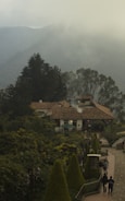 A rustic building with a tiled roof nestled among lush greenery. Smoke rises gently from the chimney, adding to the serene atmosphere. The foreground features a neatly arranged garden with trimmed bushes along a paved walkway. In the distance, misty mountains provide a tranquil backdrop, creating a picturesque setting.