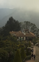 A rustic building with a tiled roof nestled among lush greenery. Smoke rises gently from the chimney, adding to the serene atmosphere. The foreground features a neatly arranged garden with trimmed bushes along a paved walkway. In the distance, misty mountains provide a tranquil backdrop, creating a picturesque setting.