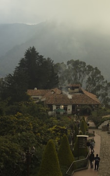 A rustic building with a tiled roof nestled among lush greenery. Smoke rises gently from the chimney, adding to the serene atmosphere. The foreground features a neatly arranged garden with trimmed bushes along a paved walkway. In the distance, misty mountains provide a tranquil backdrop, creating a picturesque setting.