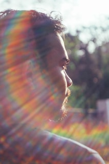 A person is seen in profile with the focus on their face, looking towards a light source. Sunlight creates a prism of rainbow colors across the image, casting colorful lines over the person. The background is blurred, with hints of greenery, suggesting an outdoor setting.