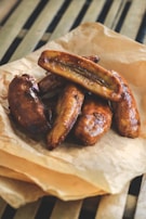 A pile of golden-brown, fried bananas resting on a crumpled piece of brown parchment paper. The bananas appear caramelized and shiny, showcasing a rich, glossy texture. The setting includes a background with wooden slats, suggesting a rustic or homemade presentation.