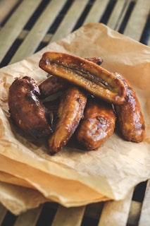 A pile of golden-brown, fried bananas resting on a crumpled piece of brown parchment paper. The bananas appear caramelized and shiny, showcasing a rich, glossy texture. The setting includes a background with wooden slats, suggesting a rustic or homemade presentation.