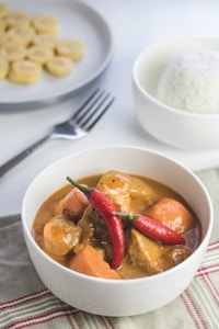 A white bowl contains a savory dish with chunks of meat and vegetables in a rich, creamy sauce, garnished with two whole red chili peppers. Nearby, a bowl of steamed white rice and a plate with slices of banana can be seen. A fork rests on the table next to the bowls. The setting is casual and inviting, placed on a striped cloth.
