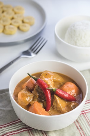 A white bowl contains a savory dish with chunks of meat and vegetables in a rich, creamy sauce, garnished with two whole red chili peppers. Nearby, a bowl of steamed white rice and a plate with slices of banana can be seen. A fork rests on the table next to the bowls. The setting is casual and inviting, placed on a striped cloth.