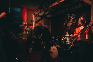 A band performing energetically in a dimly lit venue. The musicians are playing electric guitars and a drum set, with long hair moving as they play passionately. The atmosphere is intimate, with low lighting and a rustic decor, featuring wooden elements and a large decorative wall piece in the background.