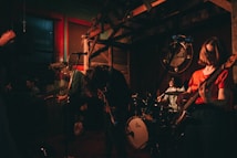 A band performing energetically in a dimly lit venue. The musicians are playing electric guitars and a drum set, with long hair moving as they play passionately. The atmosphere is intimate, with low lighting and a rustic decor, featuring wooden elements and a large decorative wall piece in the background.