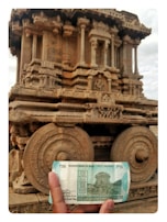 A hand is holding a 50 rupee Indian banknote in front of an intricately carved stone chariot at a historical site. The chariot features detailed sculptures and ornate designs, with large stone wheels and pillars.
