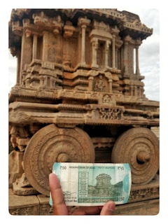 A hand is holding a 50 rupee Indian banknote in front of an intricately carved stone chariot at a historical site. The chariot features detailed sculptures and ornate designs, with large stone wheels and pillars.