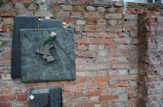 A textured brick wall features a commemorative plaque with a bronze relief sculpture depicting a map. A single white rose is placed on the plaque, possibly signifying remembrance or tribute. Below the plaque, there is a black inscribed tablet with text, further emphasizing its commemorative nature.