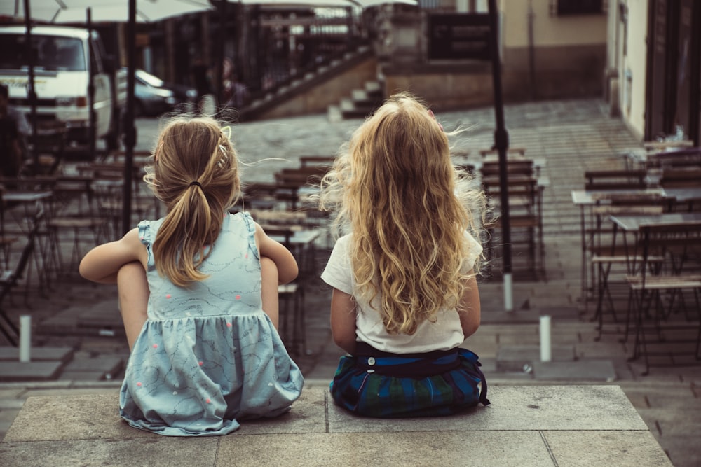 two girls sitting on concrete pavement facing on dining tables during daytime