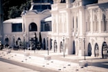 An elegant, historic building with multiple arched windows and decorative architectural details. The facade is light-colored, with tall street lamps lining the walkway in front. There's a hint of greenery in the surroundings, contributing to a serene and stately atmosphere.