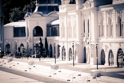 An elegant, historic building with multiple arched windows and decorative architectural details. The facade is light-colored, with tall street lamps lining the walkway in front. There's a hint of greenery in the surroundings, contributing to a serene and stately atmosphere.