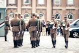A group of uniformed soldiers is performing a ceremonial march in a cobblestone square. They wear brown uniforms with green accents and carry rifles with bayonets. The scene is set in front of a historical building with large windows in the background, and several onlookers watch the procession.