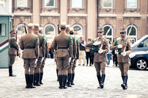 A group of uniformed soldiers is performing a ceremonial march in a cobblestone square. They wear brown uniforms with green accents and carry rifles with bayonets. The scene is set in front of a historical building with large windows in the background, and several onlookers watch the procession.