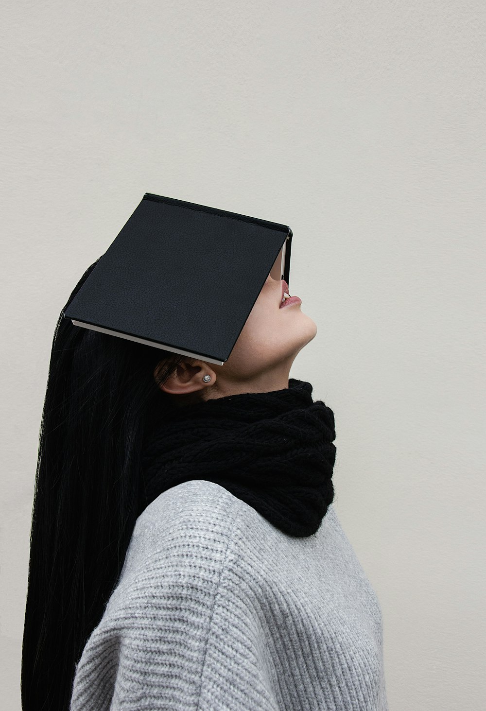 woman in gray sweater and black scarf covering face with black book