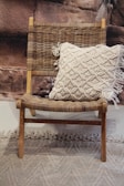 A wooden chair with a woven seat and backrest is adorned with a cream-colored, textured pillow featuring a tassel design. The backdrop consists of a rocky textured wall, and there is a textured carpet on the floor.