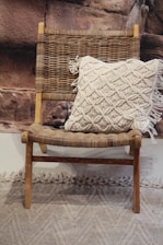 A wooden chair with a woven seat and backrest is adorned with a cream-colored, textured pillow featuring a tassel design. The backdrop consists of a rocky textured wall, and there is a textured carpet on the floor.