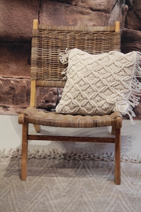 A wooden chair with a woven seat and backrest is adorned with a cream-colored, textured pillow featuring a tassel design. The backdrop consists of a rocky textured wall, and there is a textured carpet on the floor.