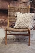 A wooden chair with a woven seat and backrest is adorned with a cream-colored, textured pillow featuring a tassel design. The backdrop consists of a rocky textured wall, and there is a textured carpet on the floor.