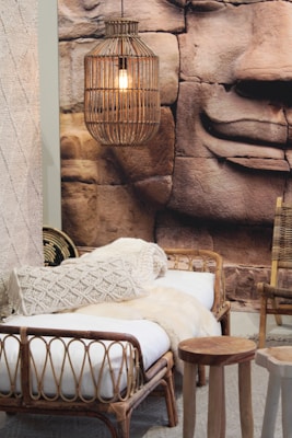 A cozy interior setup features a wicker bed with white and cream knitted blankets. A wicker pendant light hangs from above, casting a warm glow. The backdrop includes a large image of a stone sculpture with serene facial features, enhancing the natural and rustic aesthetic. A small wooden stool and a woven basket are also part of the decor.