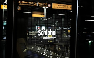 The image showcases signage for Schiphol Amsterdam Airport, prominently displayed at an airport terminal with visible glass walls and overhead lights. There is a reflective surface in the foreground, possibly a window or a screen, and illuminated signs in different languages can be seen above the main sign.