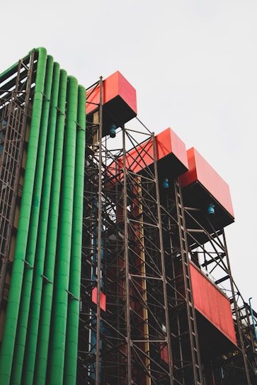 A building facade featuring an industrial design with prominent colorful pipes. Green cylindrical pipes run vertically alongside red cuboid structures, all supported by a complex network of metallic framing. The structure's design is modern and unconventional, suggesting a blend of functionality and artistic expression.
