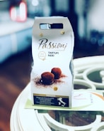 The image features a package of 'Passioni Tartufi Neri', a product that appears to be Italian truffles. The packaging is predominantly white with brown and gold accents, showcasing an image of chocolate truffles dusted with cocoa. The foreground is in focus, while the background is blurred, indicating the photo was taken in a kitchen or dining setting. There is text in Italian describing the product.