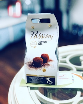 The image features a package of 'Passioni Tartufi Neri', a product that appears to be Italian truffles. The packaging is predominantly white with brown and gold accents, showcasing an image of chocolate truffles dusted with cocoa. The foreground is in focus, while the background is blurred, indicating the photo was taken in a kitchen or dining setting. There is text in Italian describing the product.