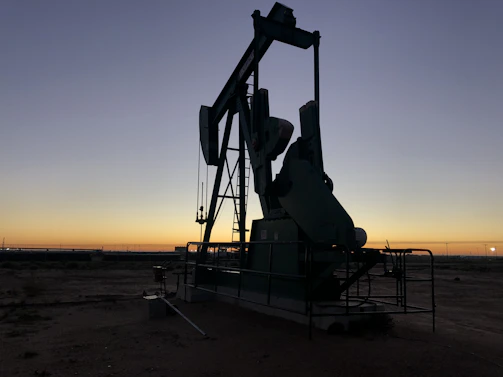 A rugged pump jack silhouetted against a sunset sky over open land.