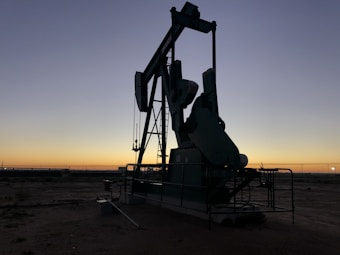 A silhouette of an oil pumpjack standing on barren land during sunset or sunrise. The sky is clear with a gradient ranging from dark blue to orange near the horizon. The machinery is prominently featured in the foreground while the expansive flat landscape stretches towards the horizon.