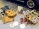 A cozy picnic setup features a guitar with a colorful design resting on a wooden bead mat, next to a blue plaid blanket. A wicker basket filled with apples, pears, and flowers sits on a striped cloth. Nearby are a red thermos, plastic plates and cutlery, and a small Fiat car advertisement. The background shows part of a blue vintage car with an intricate wheel design.
