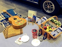 A cozy picnic setup features a guitar with a colorful design resting on a wooden bead mat, next to a blue plaid blanket. A wicker basket filled with apples, pears, and flowers sits on a striped cloth. Nearby are a red thermos, plastic plates and cutlery, and a small Fiat car advertisement. The background shows part of a blue vintage car with an intricate wheel design.