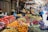 A bustling outdoor market with a variety of colorful fruits and vegetables arranged on tables. Numerous types of produce are visible, including tomatoes, oranges, apples, bananas, and eggplants. Several people interact, browsing and selecting goods, while others are engaged in conversation. The market is covered by an awning, casting shadows over the stands. Price tags are attached to some of the produce. The atmosphere is lively and vibrant.