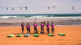 A group of people wearing wetsuits stand on the sandy beach, each with a colorful surfboard laid out in front of them. The ocean waves are visible in the background as a few surfers ride the surf. The sky is partly cloudy, adding depth to the scene. A wire with hooks runs across the top of the image.