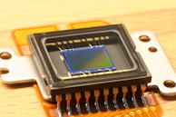 A close-up view of an electronic sensor, featuring a central square sensor surrounded by a casing and mounted on an orange circuit board. The sensor has a reflective, metallic surface with micro-components visible around its perimeter.