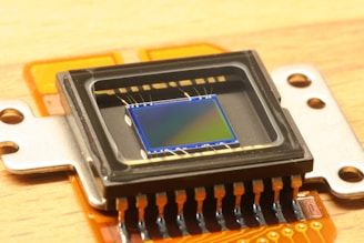 A close-up view of an electronic sensor, featuring a central square sensor surrounded by a casing and mounted on an orange circuit board. The sensor has a reflective, metallic surface with micro-components visible around its perimeter.