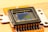 A close-up view of an electronic sensor, featuring a central square sensor surrounded by a casing and mounted on an orange circuit board. The sensor has a reflective, metallic surface with micro-components visible around its perimeter.
