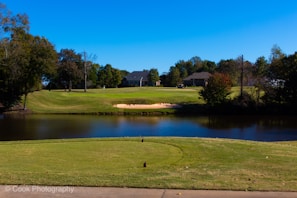 A serene golf course landscape featuring a well-maintained grassy area with a sand bunker in the middle ground. A small pond reflects the clear blue sky, bordered by lush trees. Two houses are visible in the background surrounded by more greenery, creating a peaceful environment.