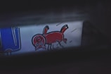 A cartoon-like drawing features a red animal with a large head and four legs, possibly depicting a pig or a whimsical creature. To the left, there is a blue object with white outlines that might resemble an abstract piece or part of a larger illustration.