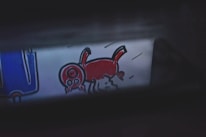 A cartoon-like drawing features a red animal with a large head and four legs, possibly depicting a pig or a whimsical creature. To the left, there is a blue object with white outlines that might resemble an abstract piece or part of a larger illustration.