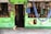 A small shop with vibrant green wooden walls displays a variety of colorful handcrafted items, including hanging ornaments and bags. A hammock is strung to the right, and a curious cat sits near the entrance, observing the scene. Woven fans, shells, and decorative masks are attached to the outer walls, adding to the eclectic charm.