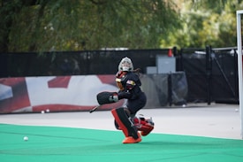 A field hockey goalkeeper wearing protective gear, including a helmet, chest pad, and leg guards, is actively positioned on a green artificial turf. The person seems focused and ready to block a ball. The background includes a blurred fence and some trees, creating an outdoor sports field setting.