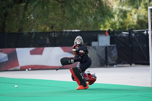 A field hockey goalkeeper wearing protective gear, including a helmet, chest pad, and leg guards, is actively positioned on a green artificial turf. The person seems focused and ready to block a ball. The background includes a blurred fence and some trees, creating an outdoor sports field setting.