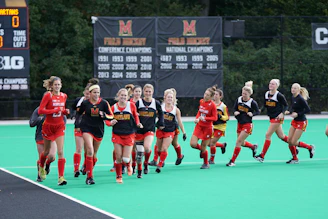 A group of female field hockey players wearing Maryland uniforms are jogging on a field. The players are in black and red uniforms, and the field is a bright green color. Multiple championship banners are displayed in the background along with a scoreboard.