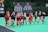A group of female field hockey players wearing Maryland uniforms are jogging on a field. The players are in black and red uniforms, and the field is a bright green color. Multiple championship banners are displayed in the background along with a scoreboard.