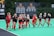 A group of female field hockey players wearing Maryland uniforms are jogging on a field. The players are in black and red uniforms, and the field is a bright green color. Multiple championship banners are displayed in the background along with a scoreboard.